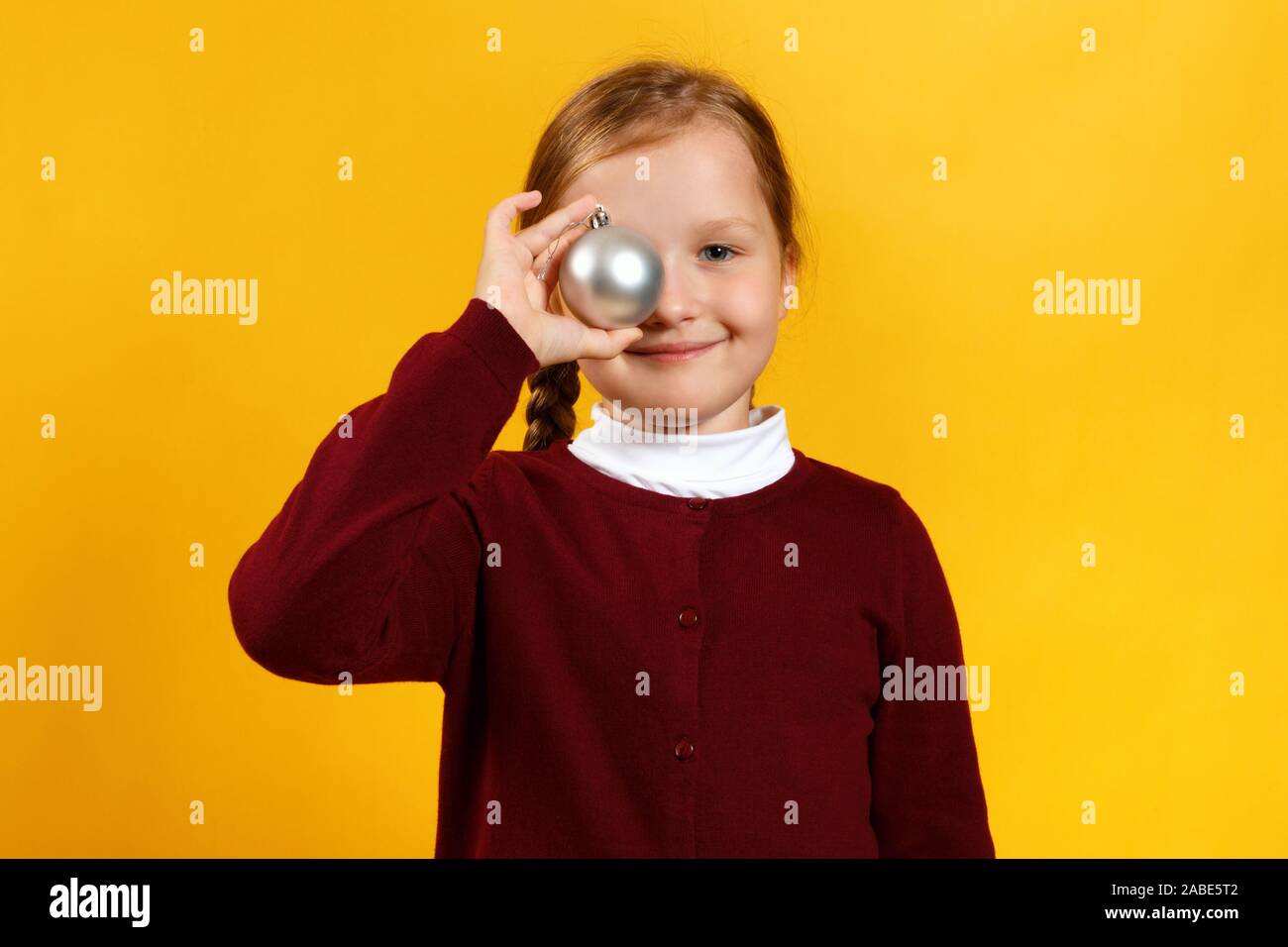 Carino piccolo felice ragazza con palla di Natale giocattolo. Closeup bambino in un maglione vinica su sfondo giallo. Foto Stock