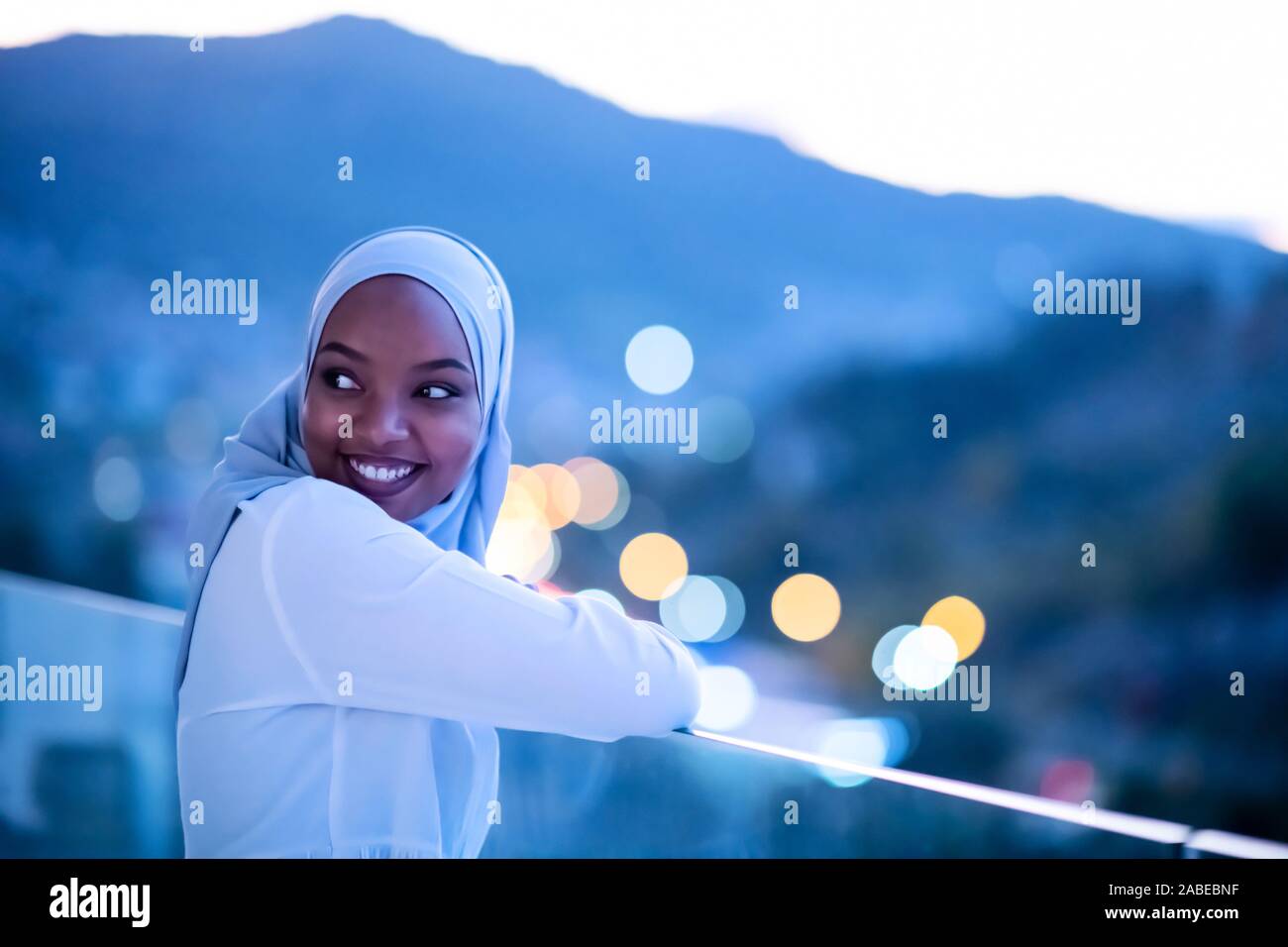 Moderna africana donna musulmana in notte al balcone sorridente in telecamera con city bokeh luci in background Foto Stock