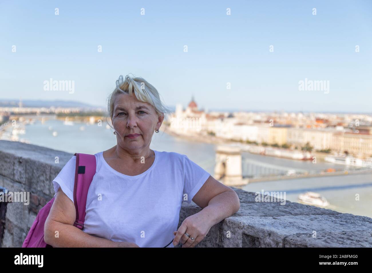 Adulto bionda donna turistico in una T-shirt bianco, con un colore rosso cremisi zaino guardare la telecamera contro lo sfondo della vecchia città di Budapest. Tr Foto Stock