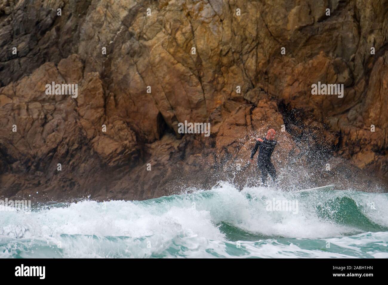 Surfer in wetsuit a cavallo di un onda sulla tavola da surf pericolosamente vicino a rocce di scogliera sul mare A la Pointe du Raz, Finistère Bretagna, Francia Foto Stock
