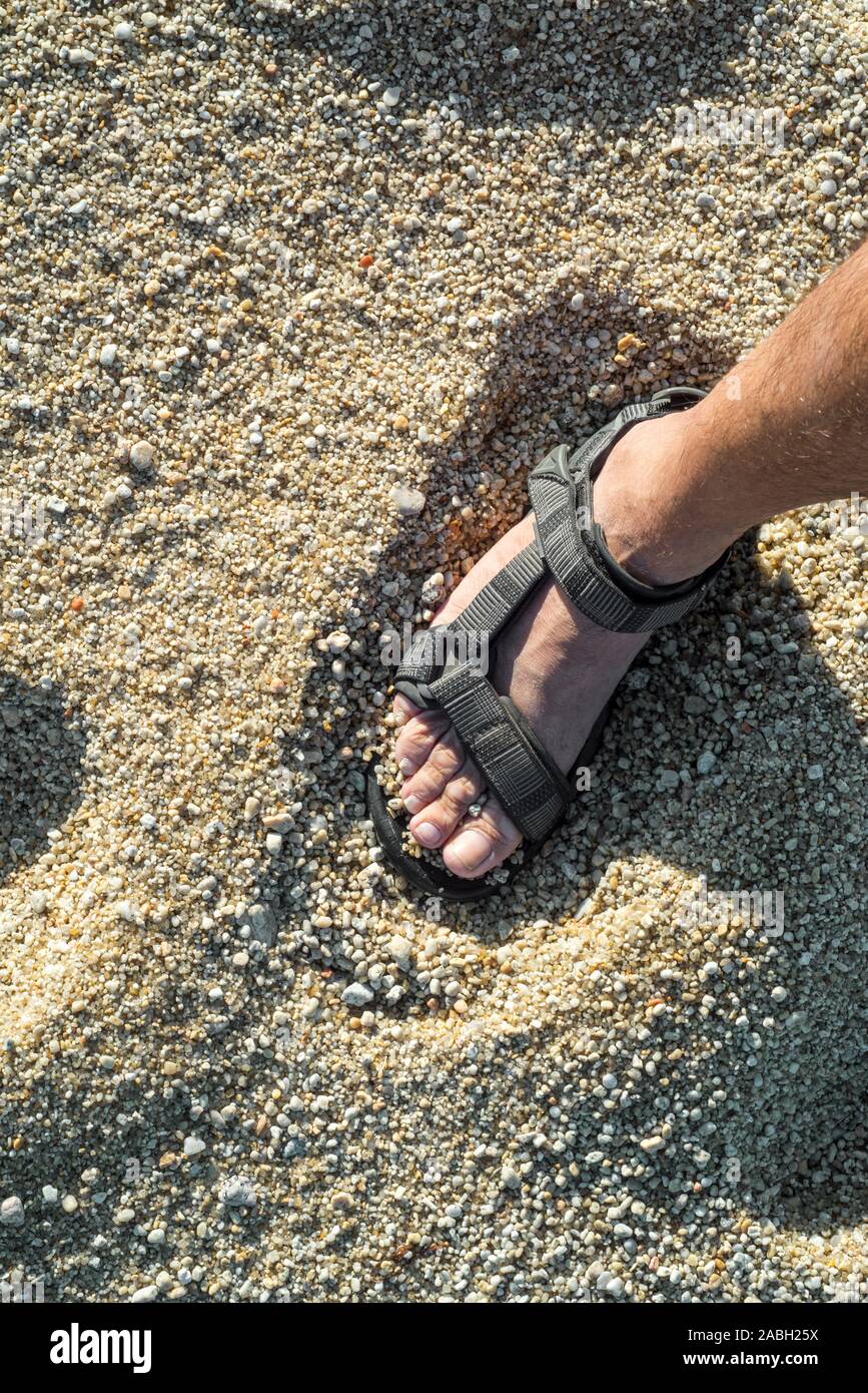 Close-up del piede maschile in sandalo permanente sulla spiaggia con mare grosso granelli di sabbia Foto Stock