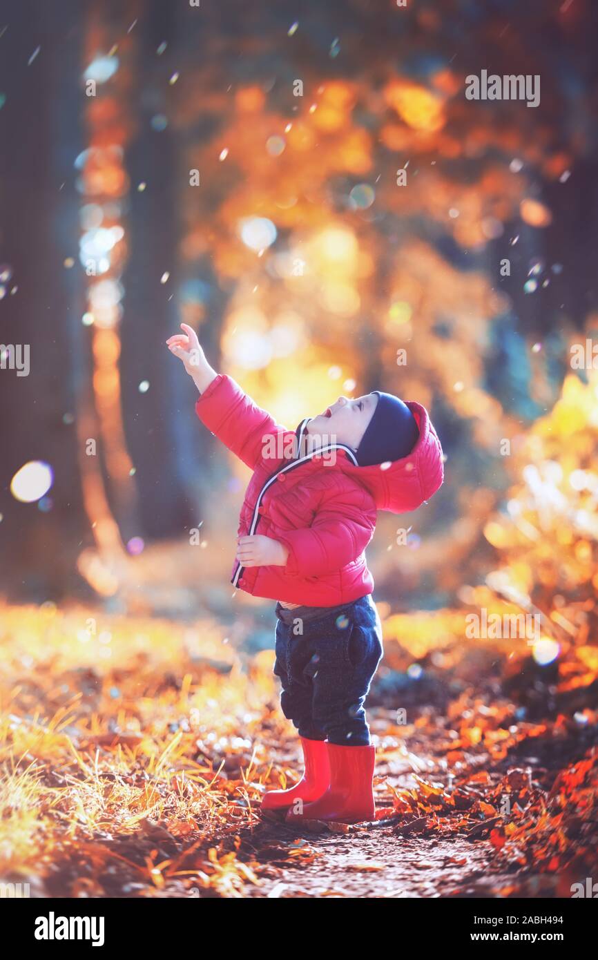 Poco toddler boy in rosso stivali di gomma e giacca rossa la cattura di gocce di pioggia in autunno park. Arancio foglie della foresta sullo sfondo Foto Stock