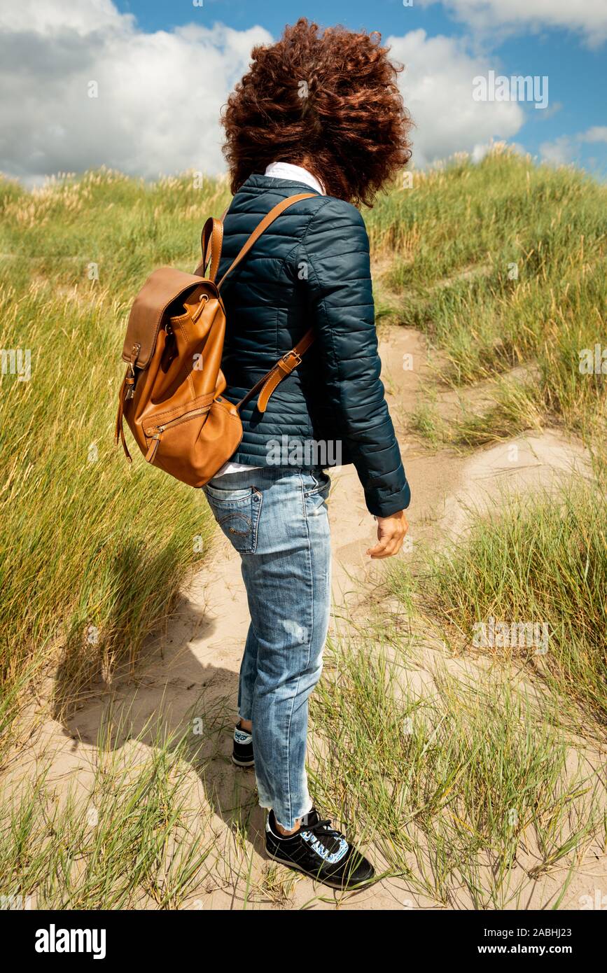 Modello rilasciato giovane donna con capelli ricci rossi indossando giacca casual e jeans blu a piedi sulla duna di sabbia costiera il giorno ventoso. Foto Stock