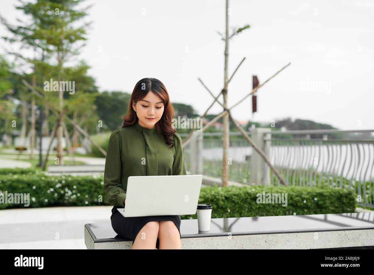 Ritratto di vendite occupato donna seduta al business park e usando il suo computer portatile durante la scrittura di testo su cellulare. Imprenditrice di lavoro on-line. Foto Stock