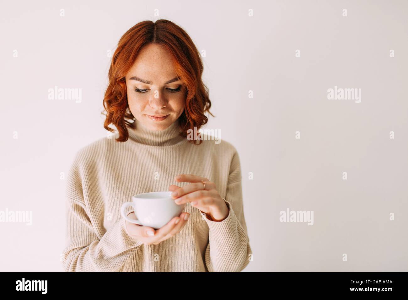 Stile di vita ritratto di dai capelli rossi bellissima ragazza con una tazza di caffè, guardando in giù a sognare. Indossa un maglione accogliente di una tonalità pastello. Foto Stock