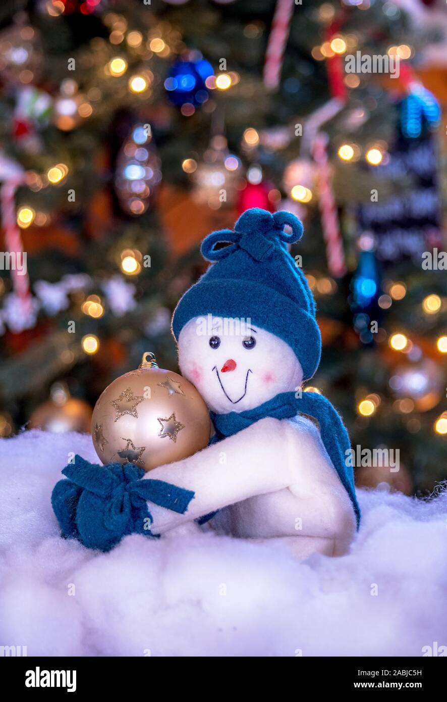 Adorabili e uomo di neve tiene un ornamento d'oro come egli si pone in un cotone snow bank da parte di un albero di Natale decorato Foto Stock