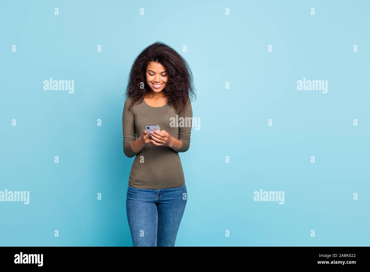 Ritratto di Allegro positivo focalizzata blogger mulatto ragazza utilizzando il suo smartphone leggere feednews tipo feedback sul blog di usura maglione verde jeans denim Foto Stock