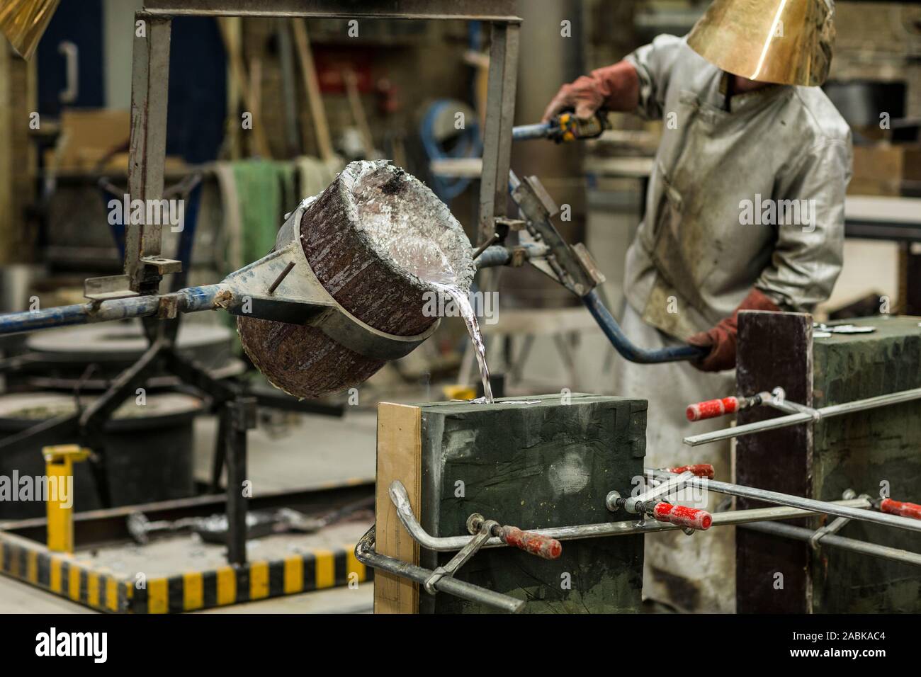 Un artigiani in argento abbigliamento protettivo di eseguire un metallo sabbia tecnica di colata con una colata di alluminio fuso colore argento liquido in uno stampo in una Foto Stock Un artigiani in argento abbigliamento protettivo di eseguire un metallo sabbia tecnica di colata con una colata di alluminio fuso colore argento liquido in uno stampo in una Foto Stock
