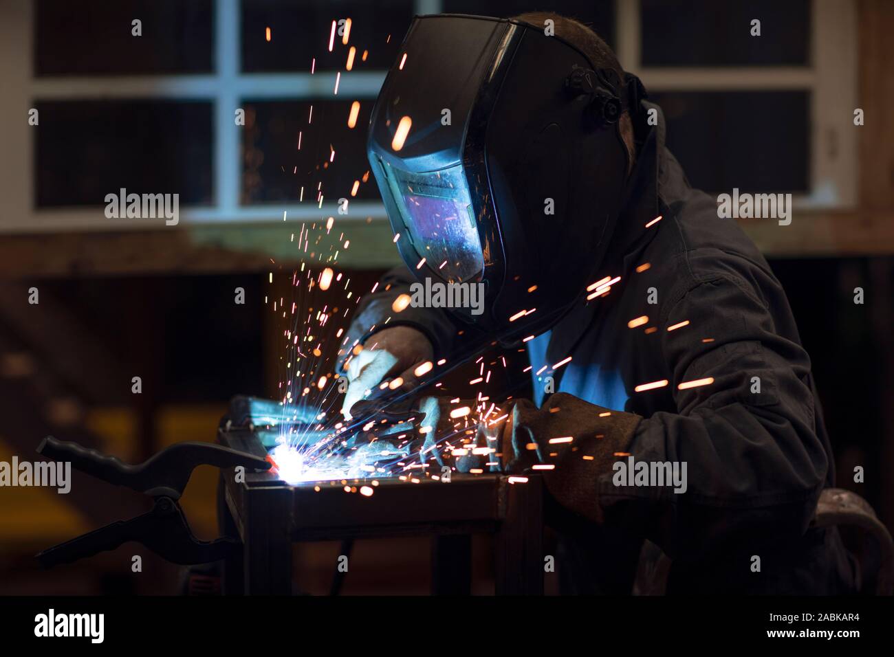 Saldatore focalizzato designer che indossa un casco e wokring su un progetto per la saldatura di pezzi di metallo mentre facendo scintille di saldatura nella sua officina di notte Foto Stock