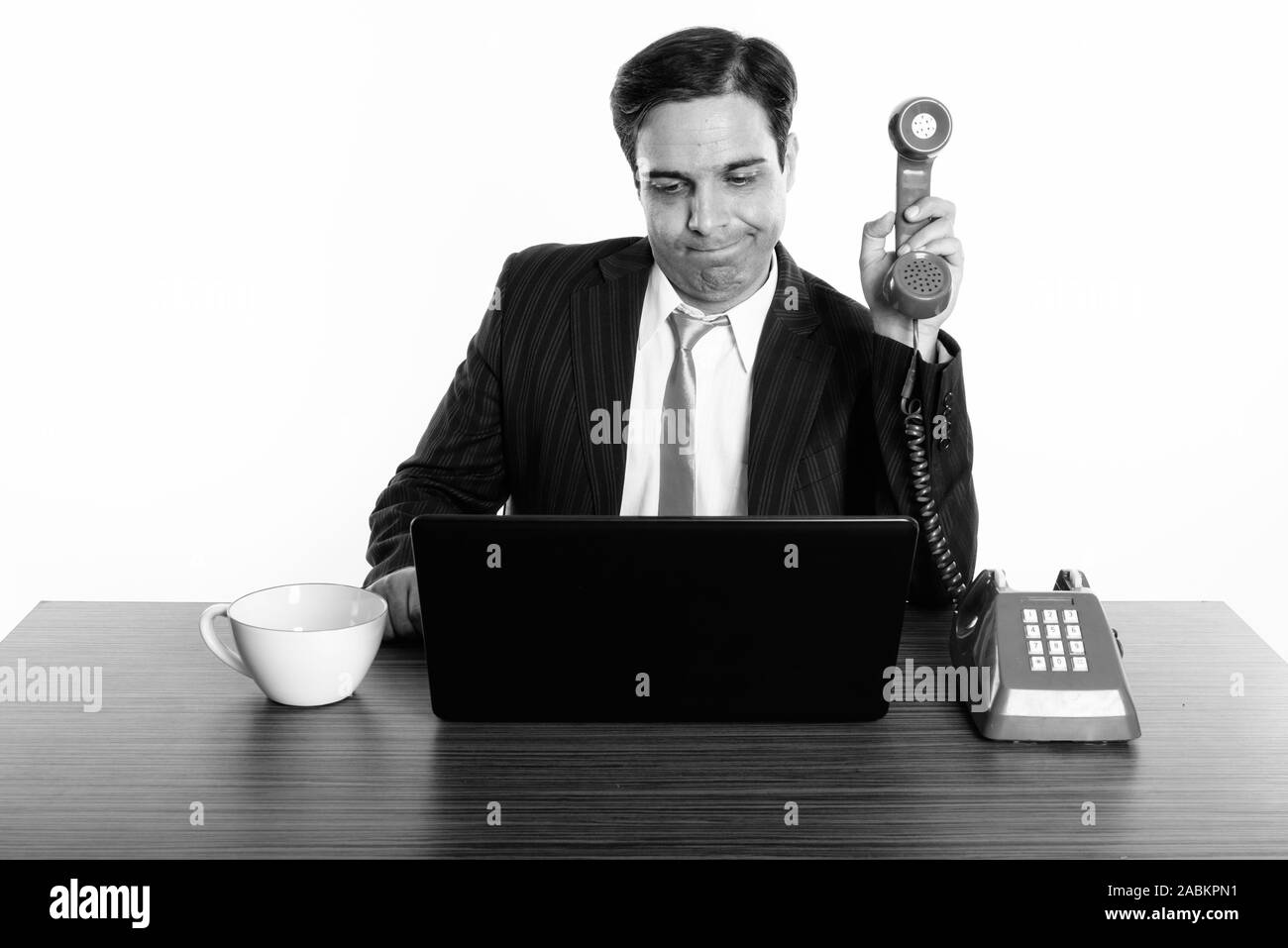 Studio shot del giovane imprenditore persiano tenendo il vecchio telefono lontano durante l'utilizzo di laptop con tazza di caffè sul tavolo in legno Foto Stock