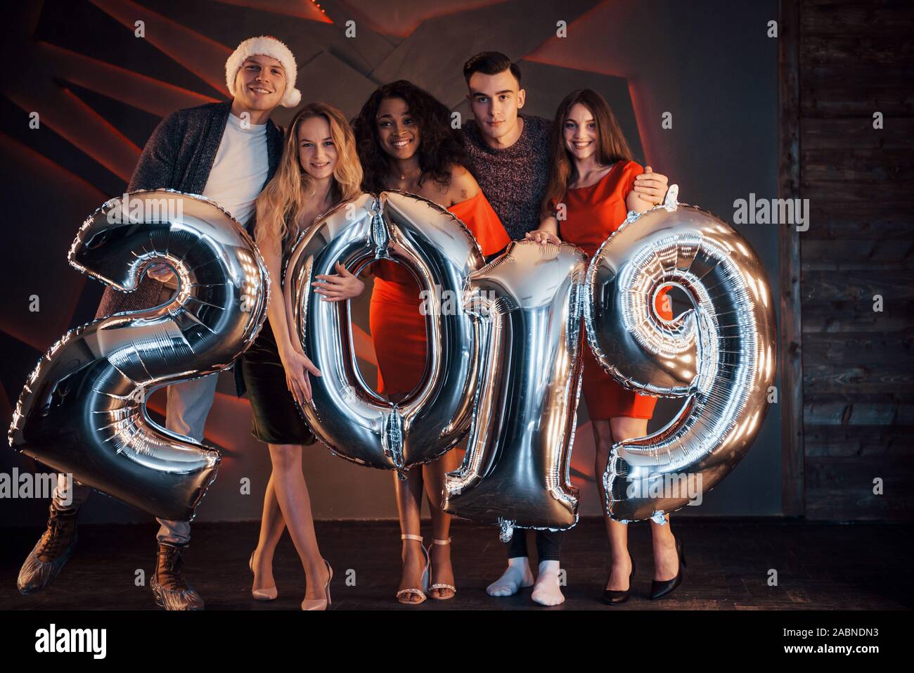 Il nuovo 2019 si avvicina. Un gruppo di giovani allegri gente multinazionale in Santa's hat con numeri di argento e gettando i confetti al partito. Felice Foto Stock