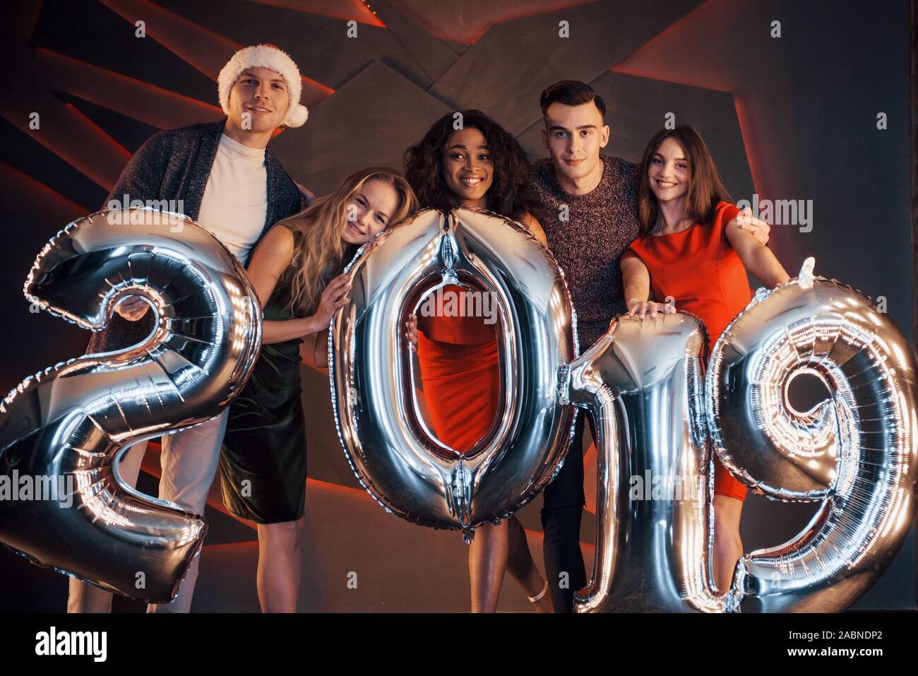 Il nuovo 2019 si avvicina. Un gruppo di giovani allegri gente multinazionale in Santa's hat con numeri di argento e gettando i confetti al partito. Felice Foto Stock