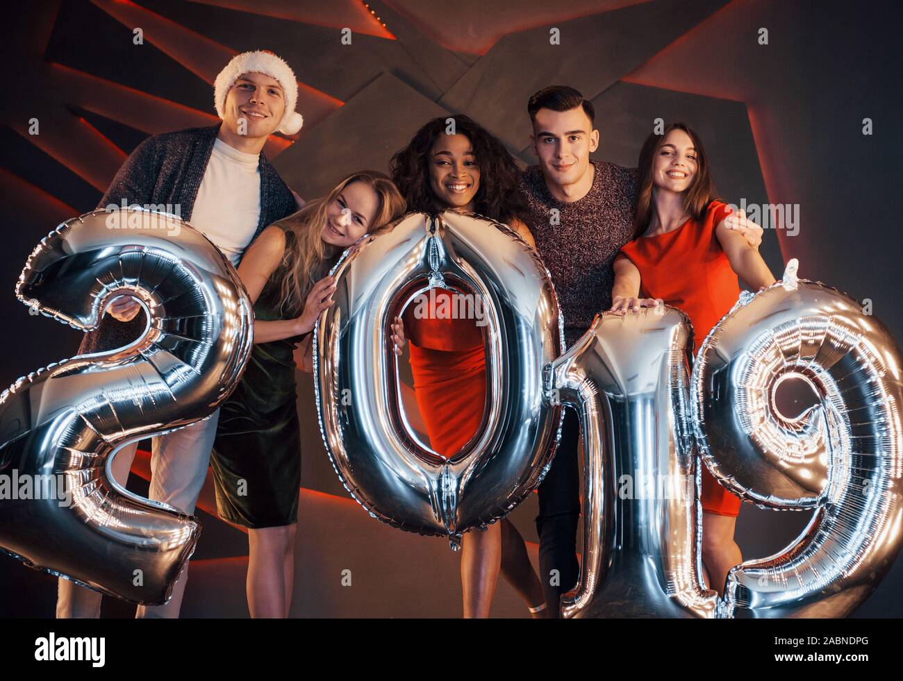 Il nuovo 2019 si avvicina. Un gruppo di giovani allegri gente multinazionale in Santa's hat con numeri di argento e gettando i confetti al partito. Felice Foto Stock