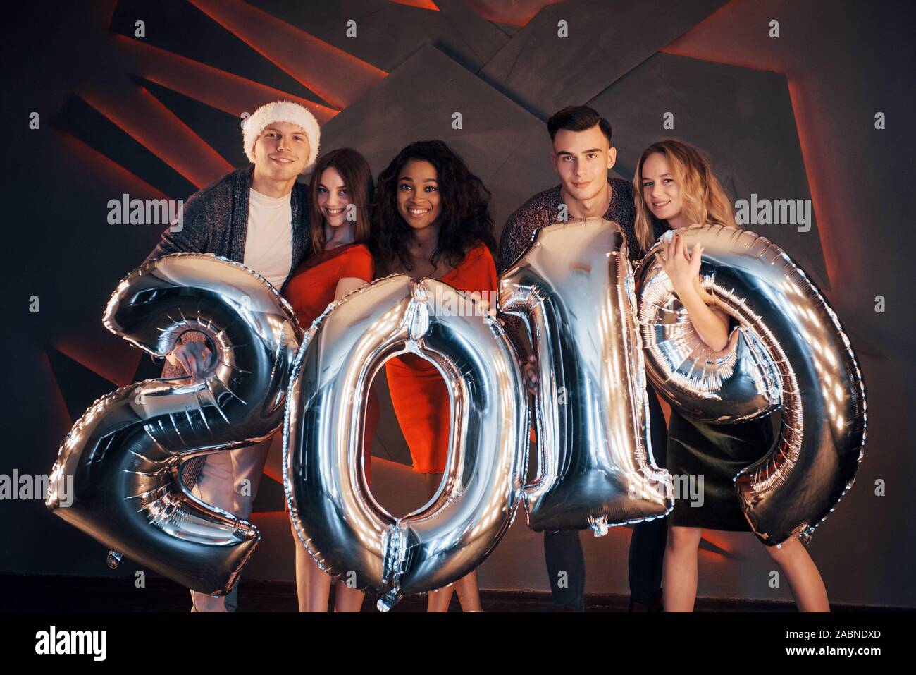 Il nuovo 2019 si avvicina. Un gruppo di giovani allegri gente multinazionale in Santa's hat con numeri di argento e gettando i confetti al partito. Felice Foto Stock