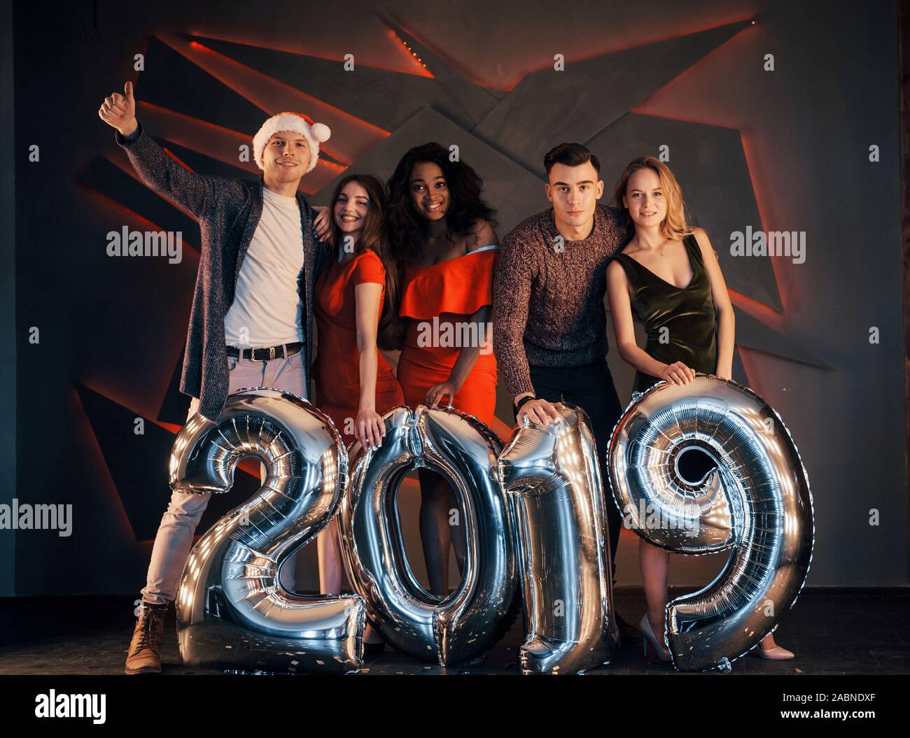 Il nuovo 2019 si avvicina. Un gruppo di giovani allegri gente multinazionale in Santa's hat con numeri di argento e gettando i confetti al partito. Felice Foto Stock