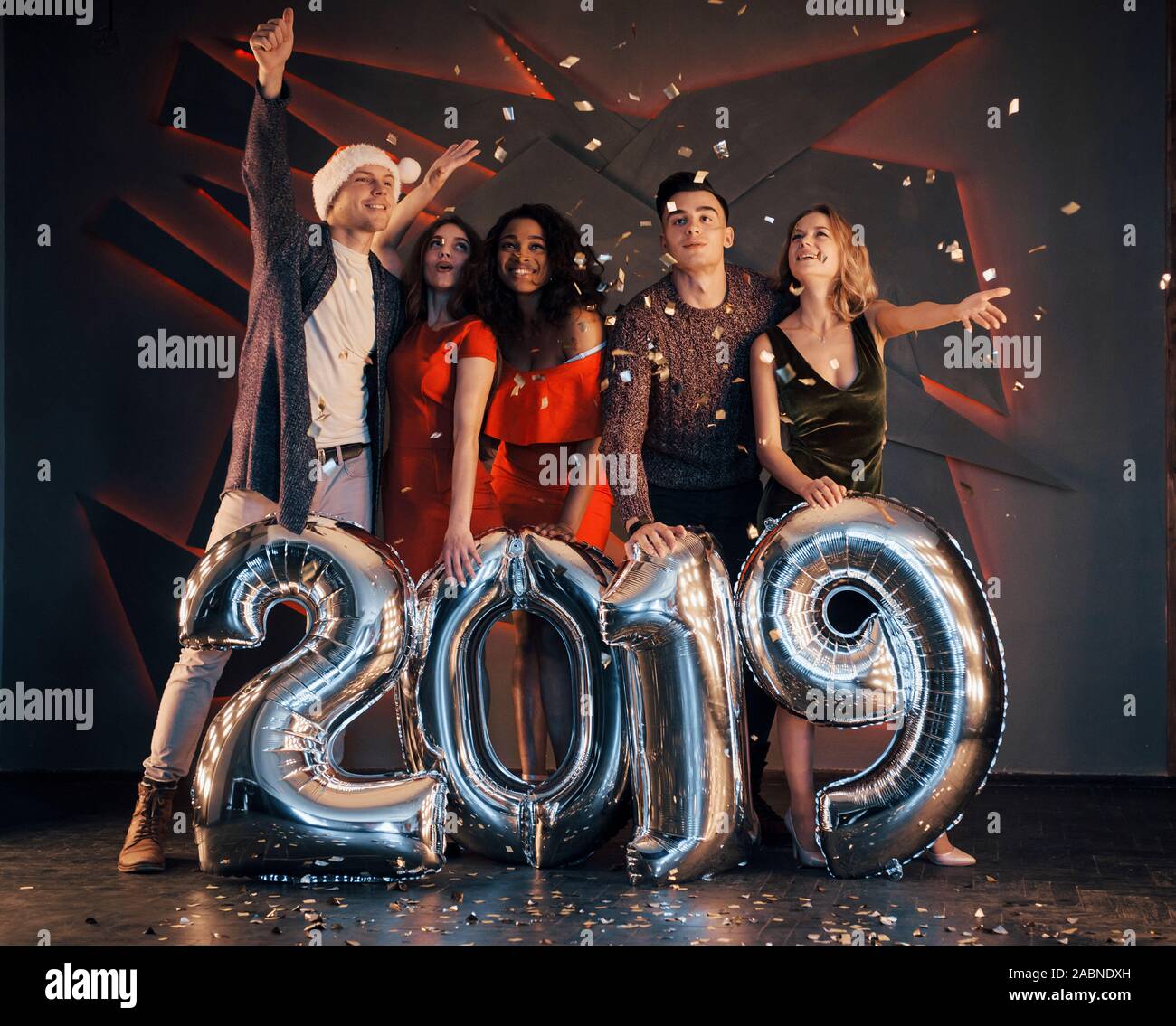 Il nuovo 2019 si avvicina. Un gruppo di giovani allegri gente multinazionale in Santa's hat con numeri di argento e gettando i confetti al partito. Felice Foto Stock