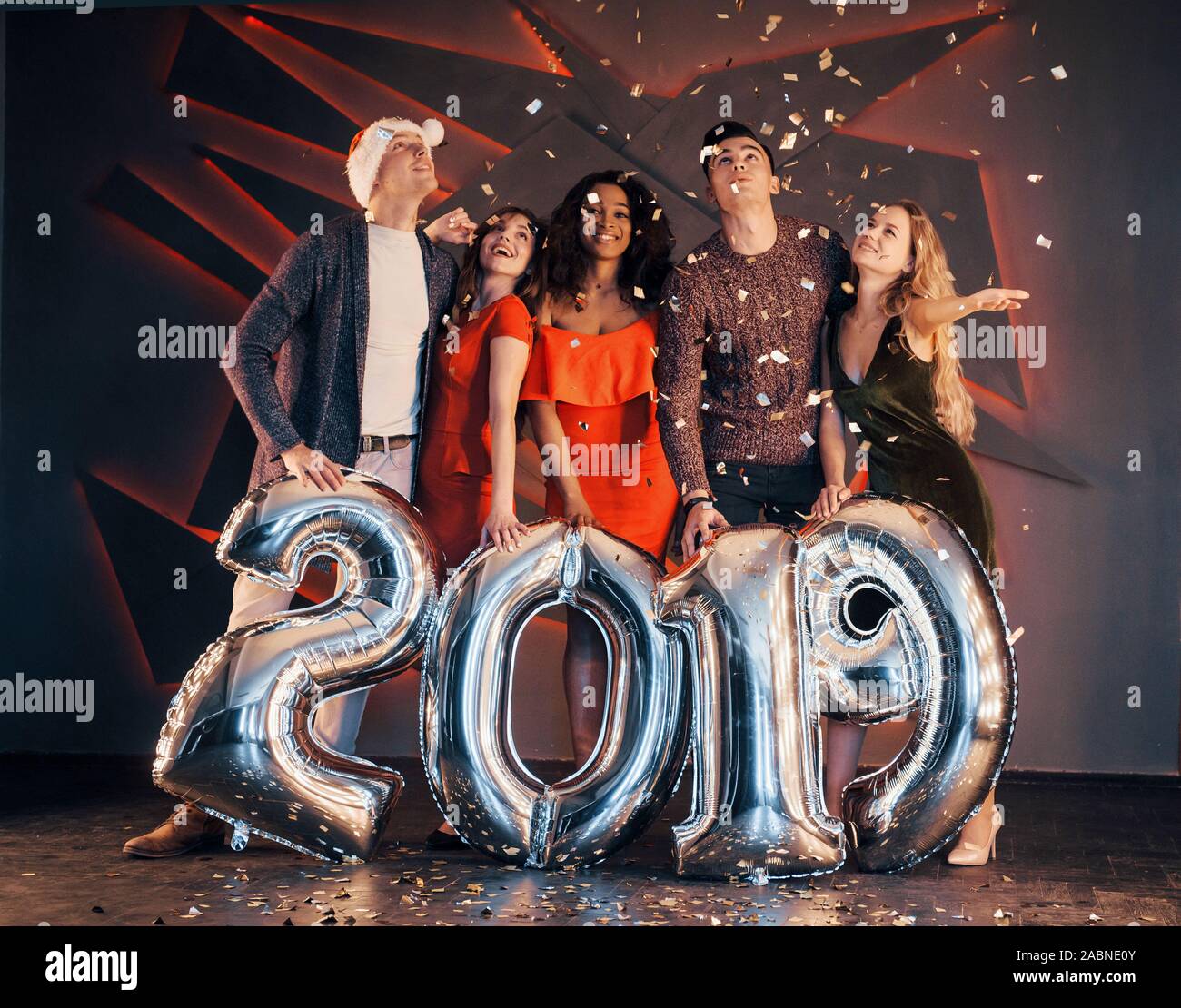 Il nuovo 2019 si avvicina. Un gruppo di giovani allegri gente multinazionale in Santa's hat con numeri di argento e gettando i confetti al partito. Felice Foto Stock