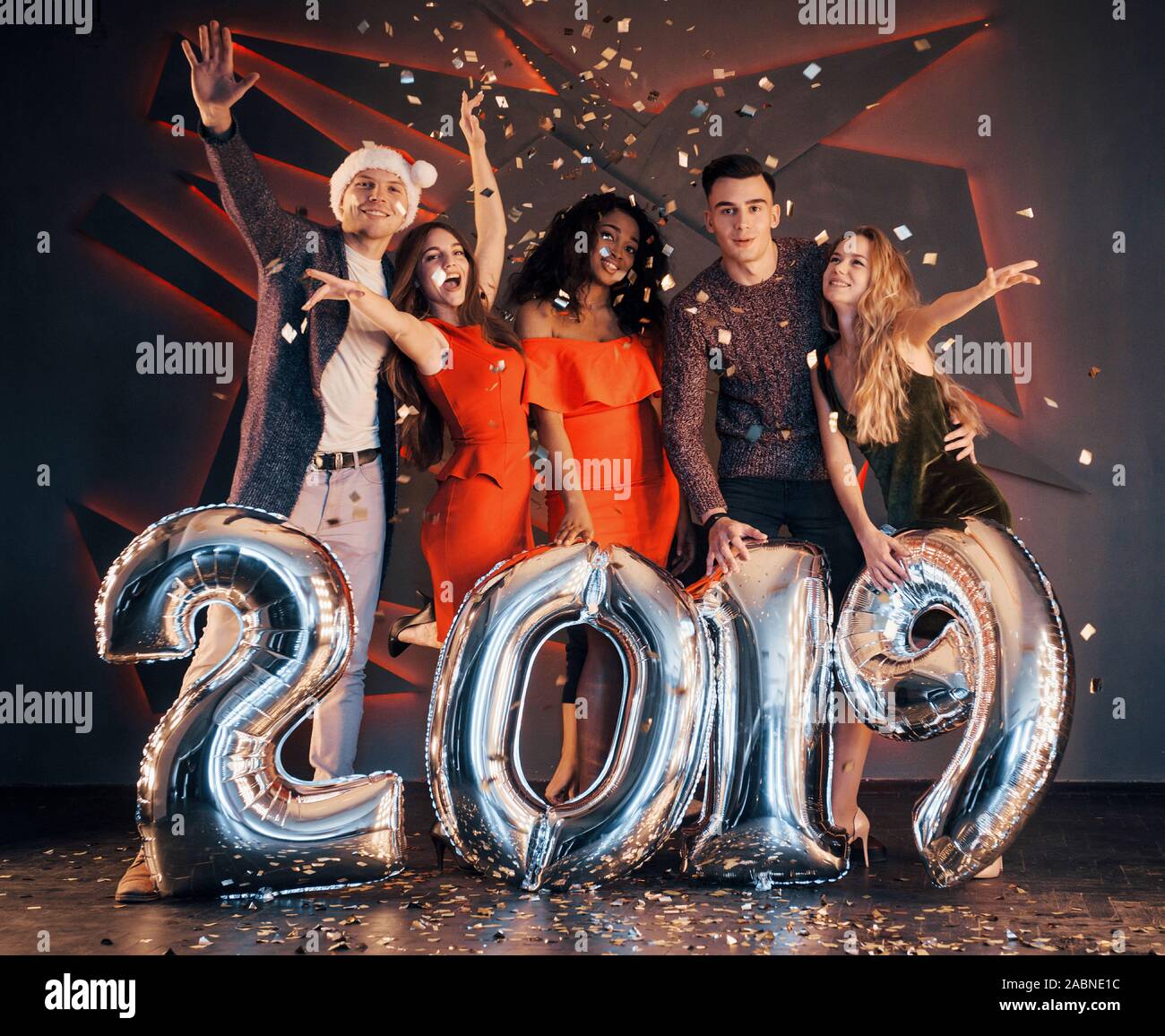 Il nuovo 2019 si avvicina. Un gruppo di giovani allegri gente multinazionale in Santa's hat con numeri di argento e gettando i confetti al partito. Felice Foto Stock