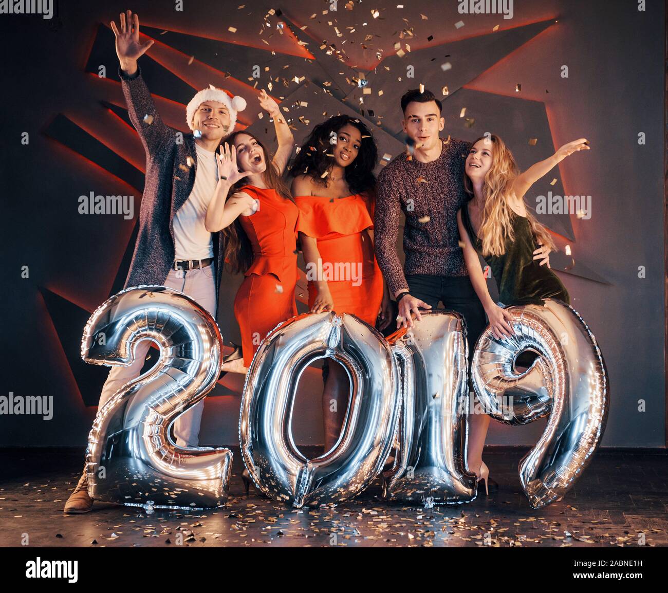 Il nuovo 2019 si avvicina. Un gruppo di giovani allegri gente multinazionale in Santa's hat con numeri di argento e gettando i confetti al partito. Felice Foto Stock