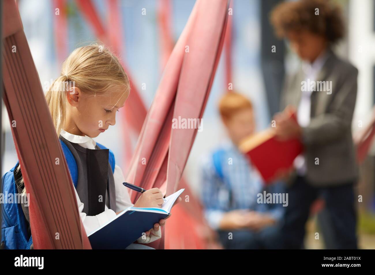 Grave schoolgirl con zaino dietro la schiena rendendo note in notebook mentre seduto sul parco giochi con i suoi compagni in background Foto Stock