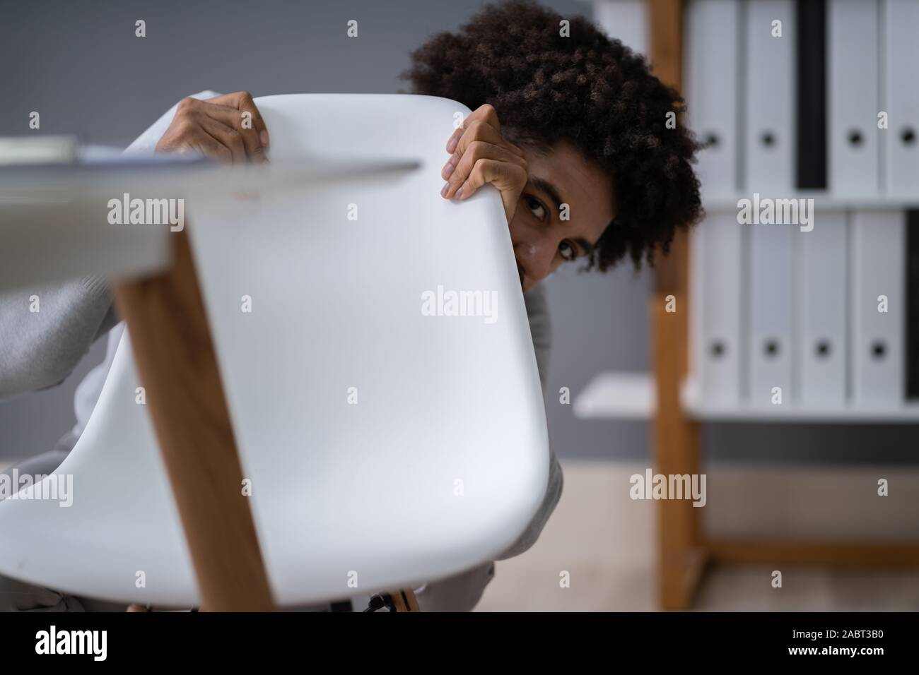 Frightened giovane imprenditore nascondersi dietro la sedia al posto di lavoro Foto Stock