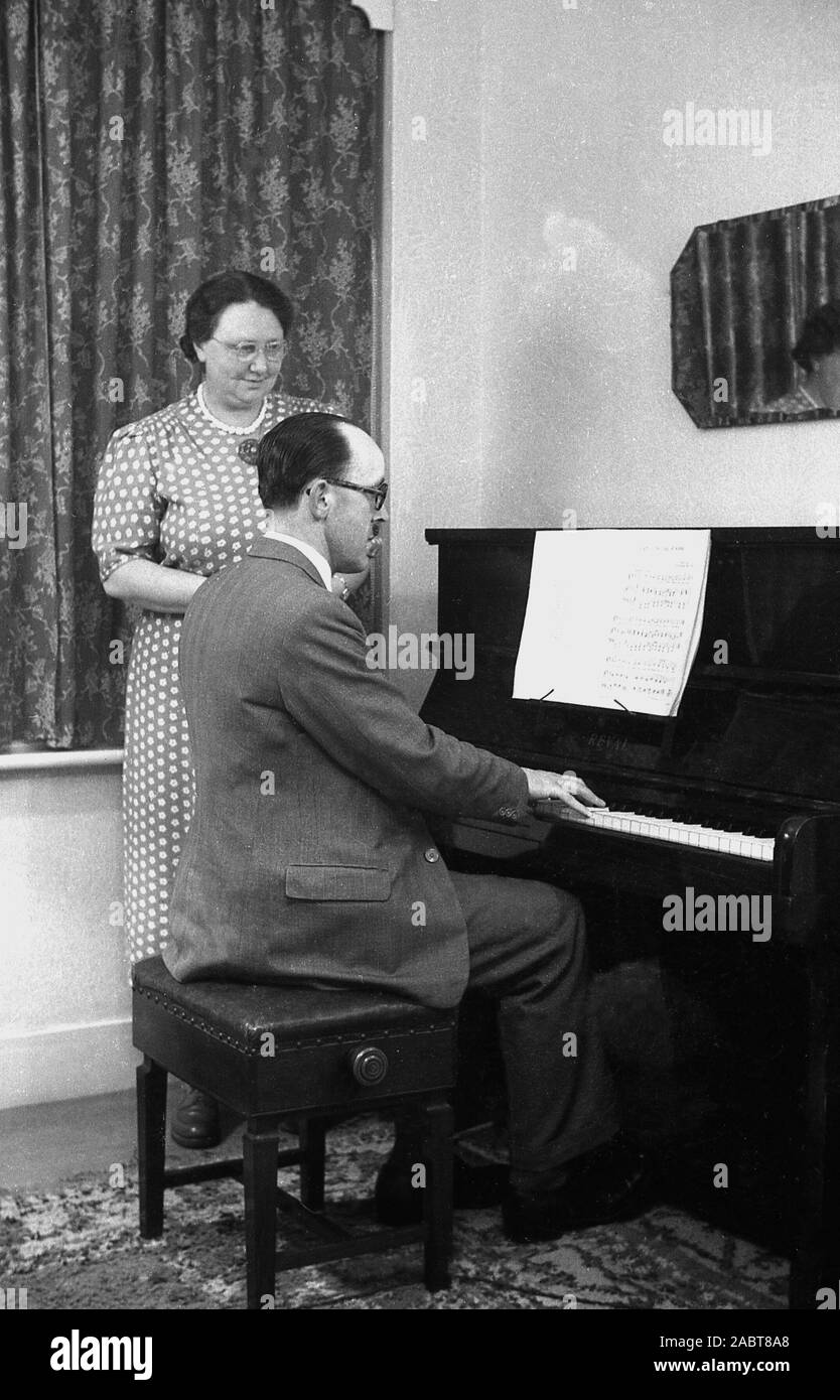 Anni '1950, storica, a casa una moglie in piedi, guardando suo marito suonare il pianoforte, a Reval, Inghilterra, Regno Unito. Nell'era prima della televisione, l'intrattenimento domestico era fornito da radio, riviste, libri e suonava strumenti musicali. Foto Stock