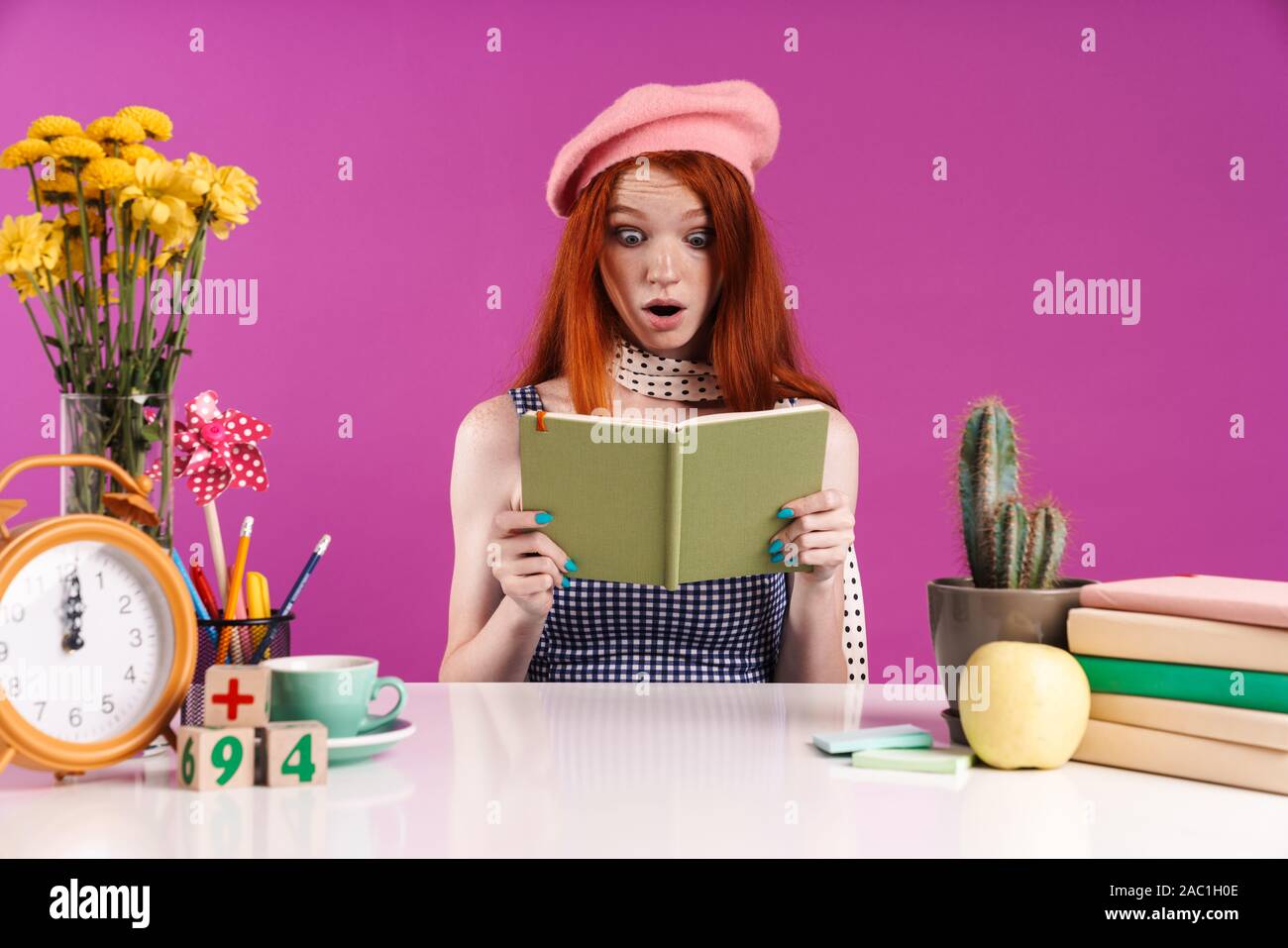 Immagine del giovane studente ragazza con diario libro mentre è seduto alla scrivania isolate su sfondo viola Foto Stock