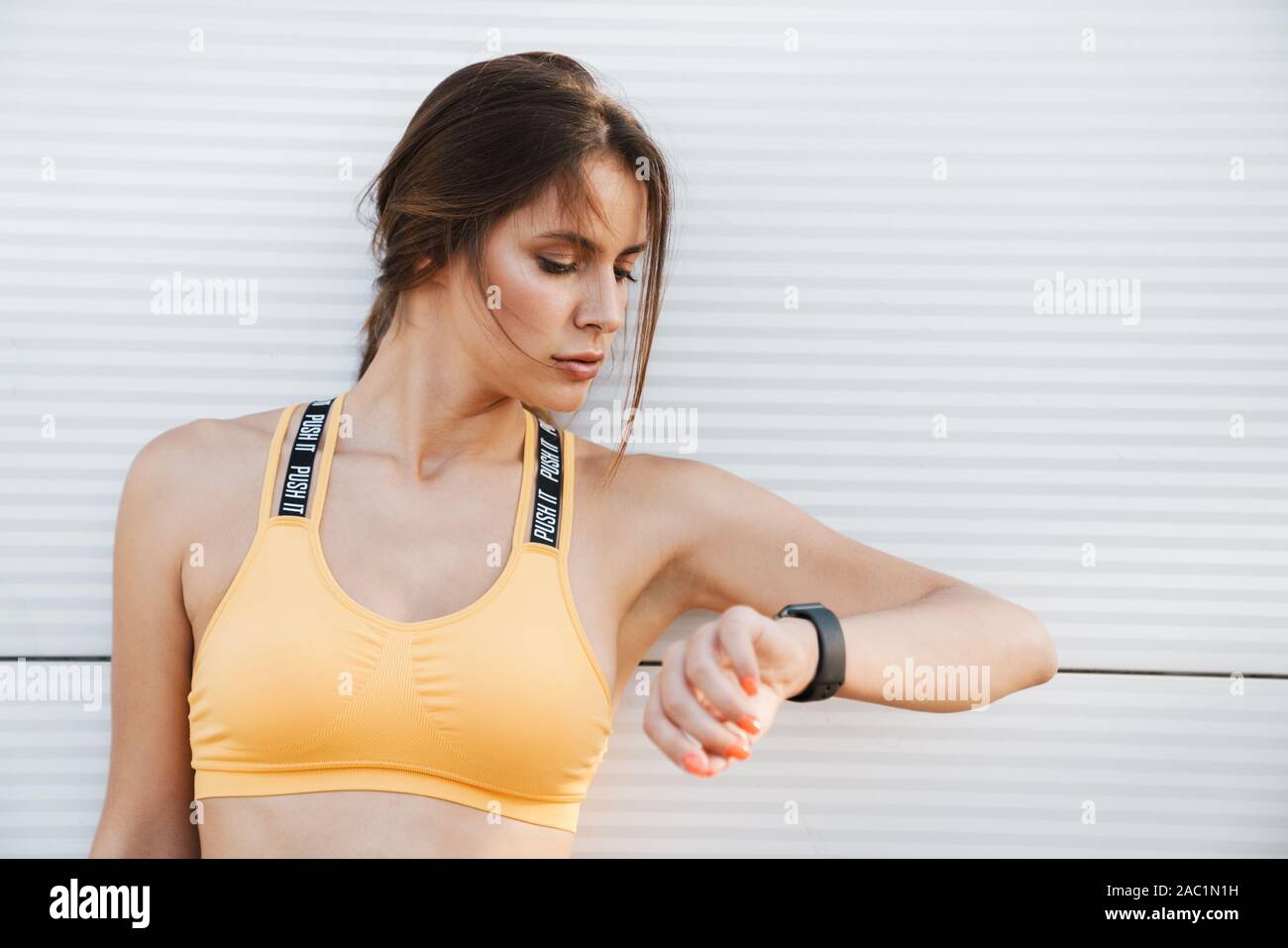 Primo piano immagine della bella donna di abbigliamento sportivo in piedi su muro bianco all'aperto e guardando un orologio da polso Foto Stock