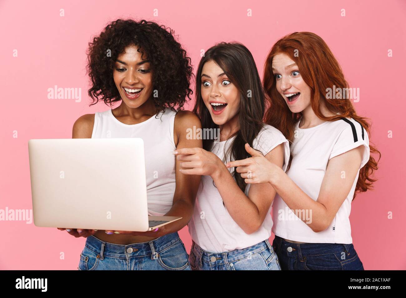 Tre Allegri ragazze multietnica in piedi isolato su sfondo rosa, puntare il dito in corrispondenza di computer portatile Foto Stock