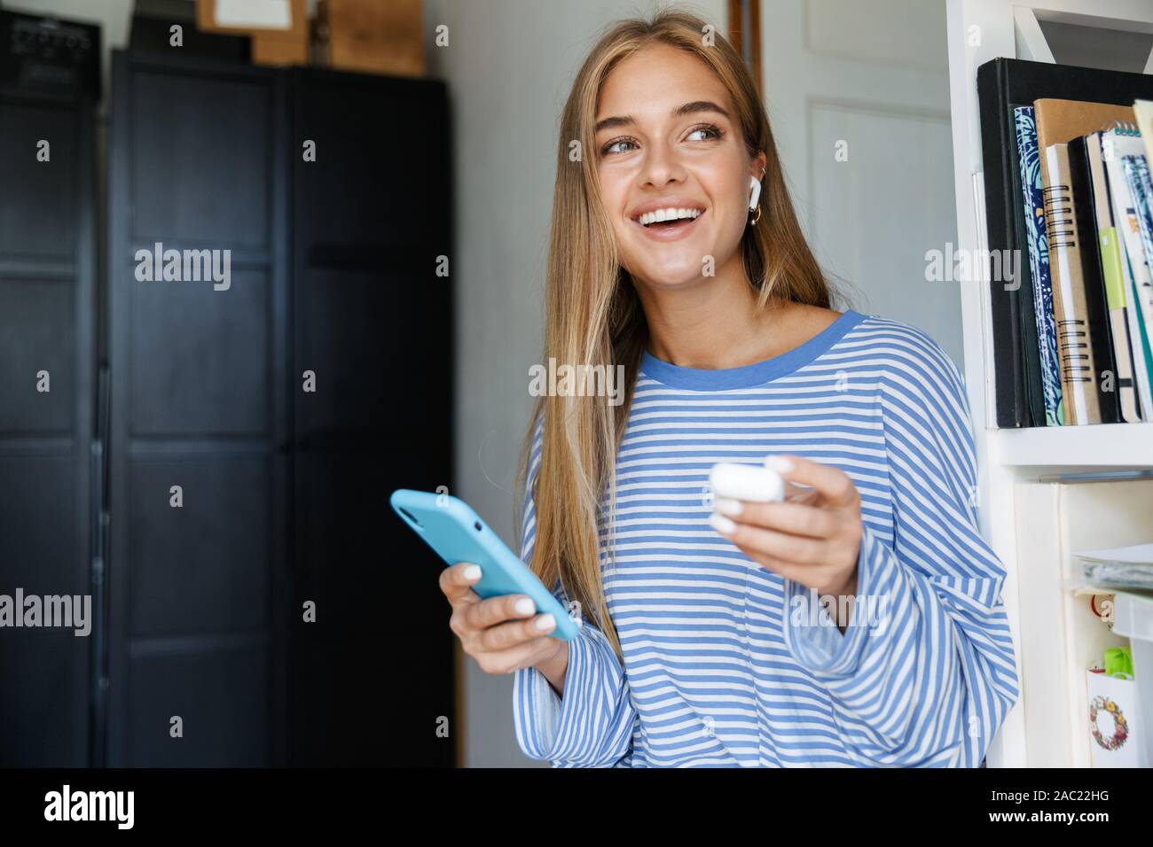 Foto di un sorridenti giovane ragazza carina a casa utilizzando il telefono cellulare mantenendo gli auricolari bluetooth. Foto Stock