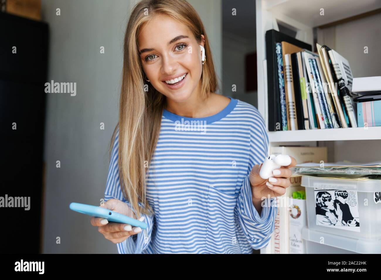 Ritratto di un ottimista giovane ragazza carina a casa utilizzando il telefono cellulare mantenendo gli auricolari bluetooth. Foto Stock