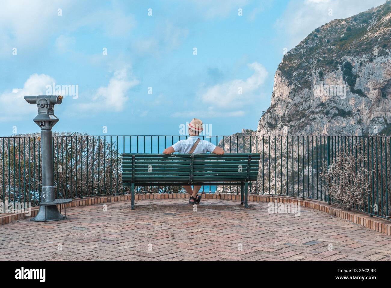Uomo che indossa hat sedersi su una panchina guardando oltre il mare Tirreno in Giardini di Augusto a Capri Foto Stock