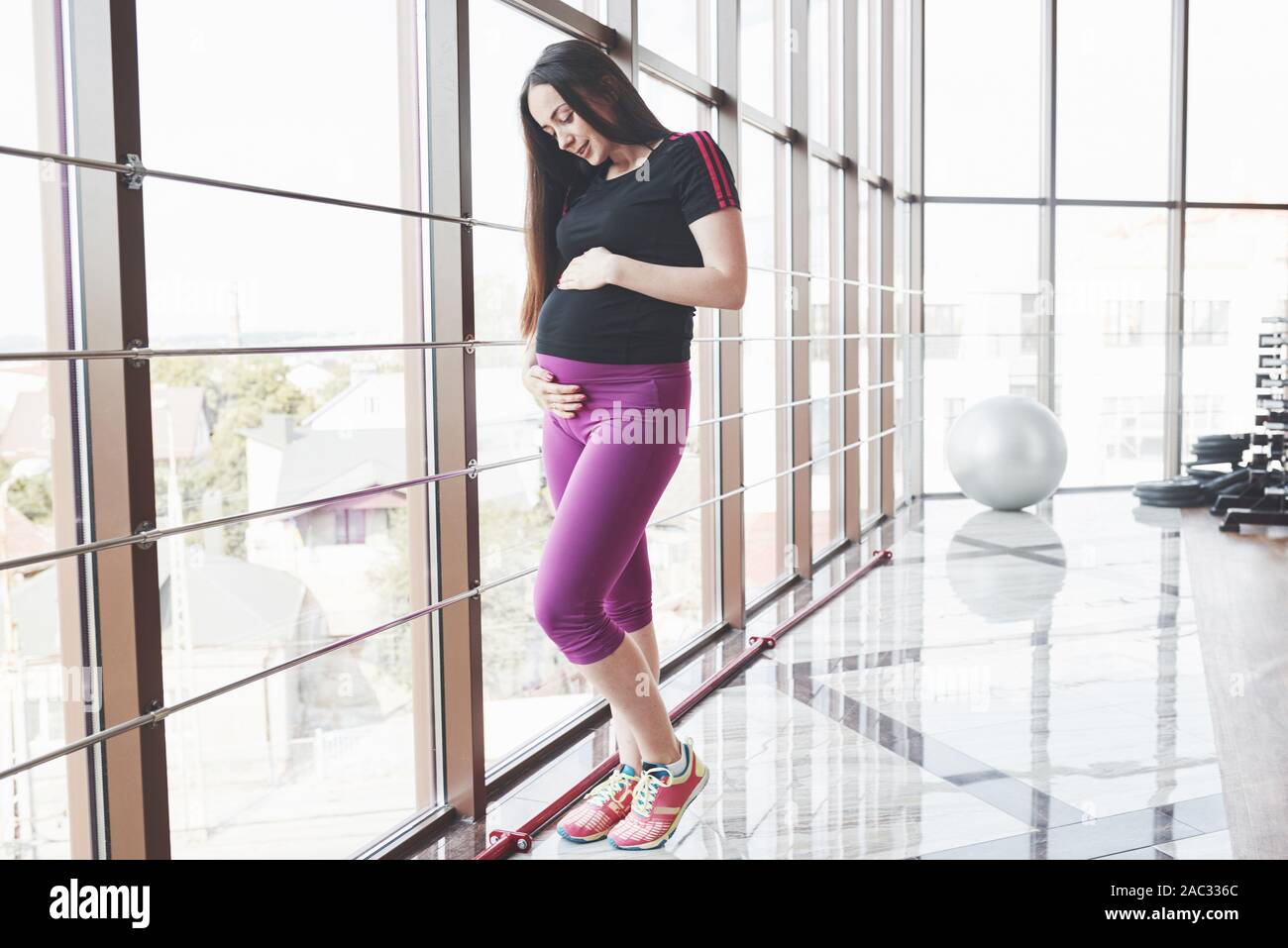Incinta brunette in piedi la palestra vicino a finestre e mantenendo le mani sulla pancia Foto Stock