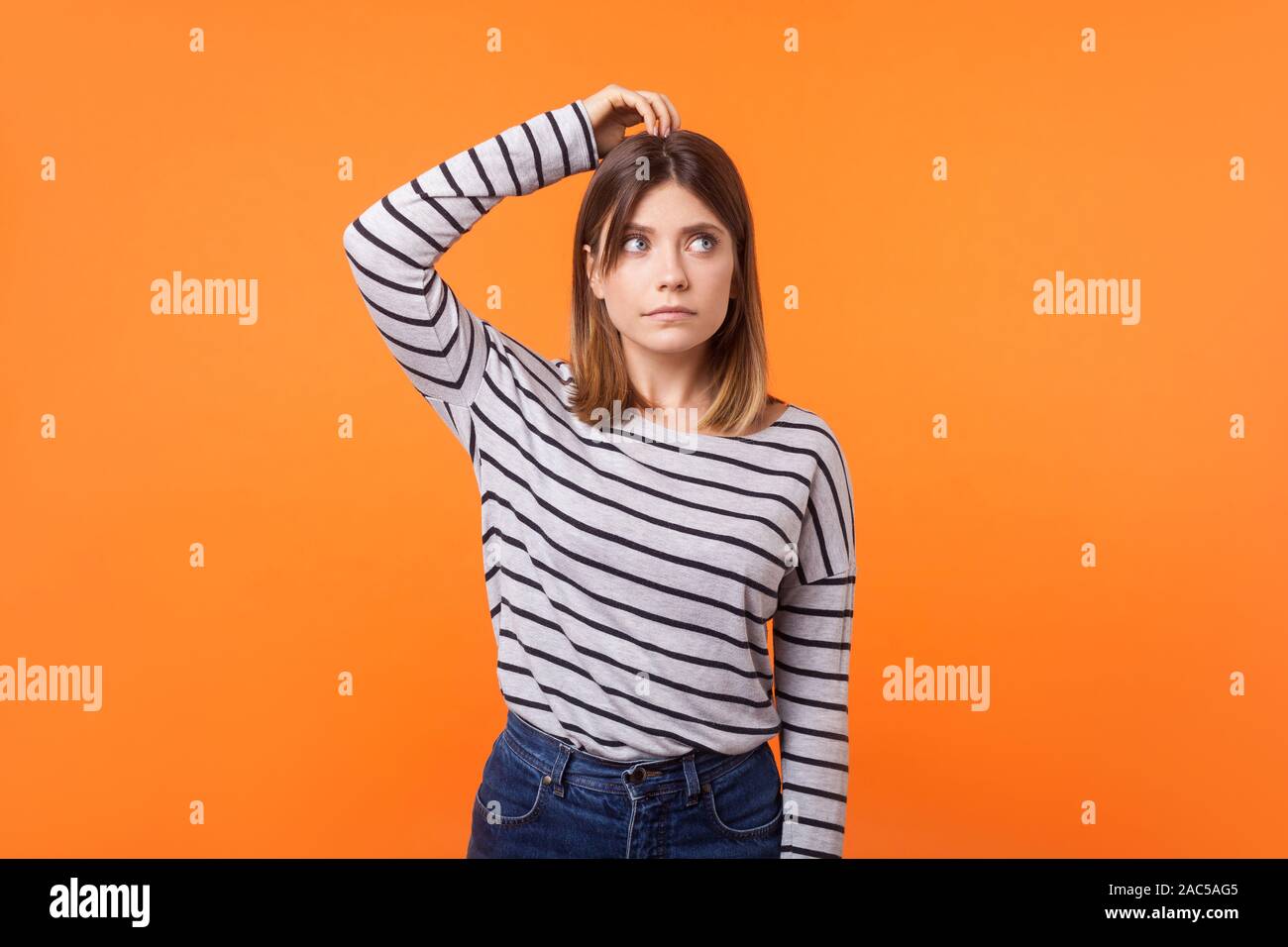 La necessità di pensare. Ritratto di confuso giovane donna con capelli castani a manica lunga camicia a righe in piedi, graffiare la testa e guardando in alto, risoluzione dei problemi Foto Stock