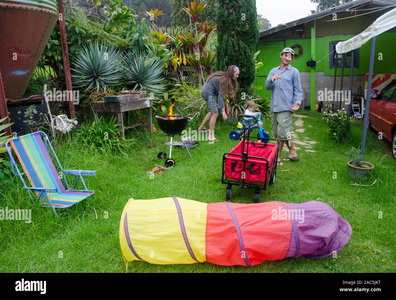 Un giovane locale madre, padre e figlio toddler giocare nel loro cortile con un barbecue a partire e giocattoli sparsi, Big Island. Foto Stock