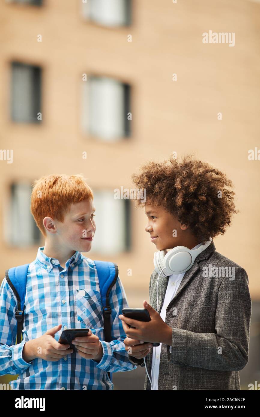 Due ragazzi sorridenti e permanente di parlarsi e utilizzando i loro smart phone all'aperto Foto Stock