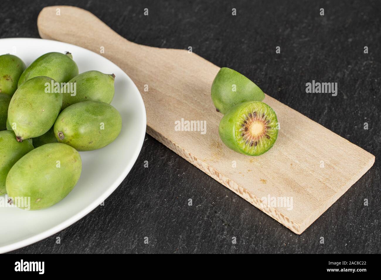 Gruppo di lotto intero di due metà di hardy kiwi verde su bianco piastra ceramica sul tagliere di legno su pietra grigia Foto Stock