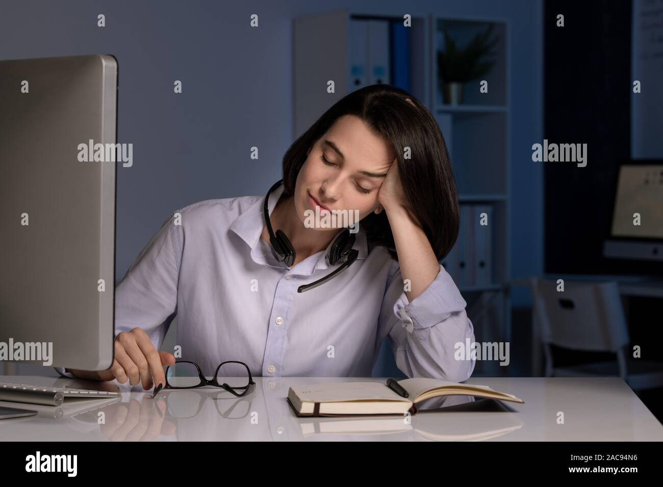 Stanco hotline femmina operatore con gli occhi chiusi napping da scrivania mentre si lavora fino a tardi la sera in ufficio scuro Foto Stock