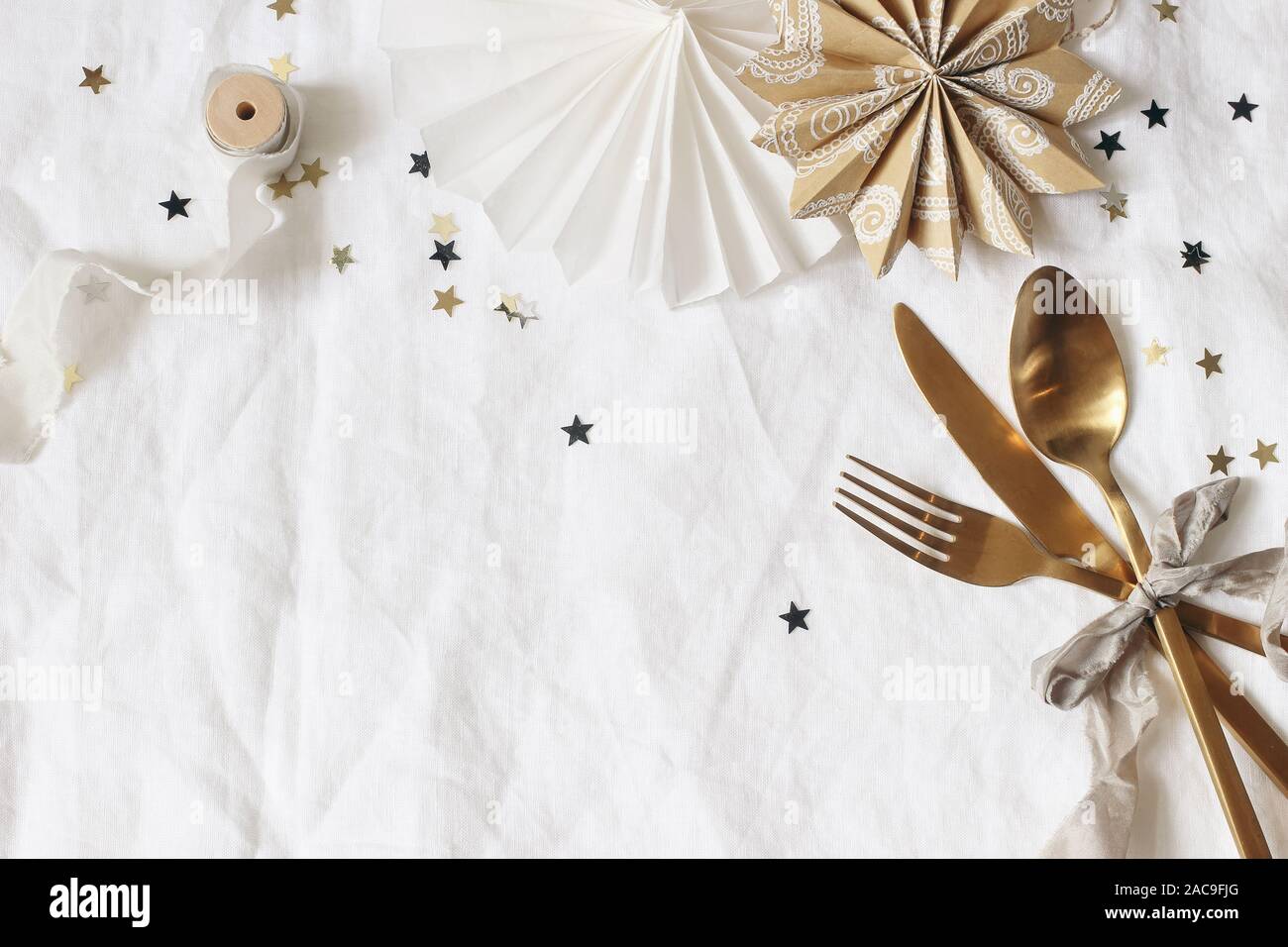 Natale e Anno Nuovo composizione. Tabella decorativo impostazione. Stelle di carta, golden posate e coriandoli stelle sulla biancheria da letto bianca dello sfondo. Celebrazione Foto Stock