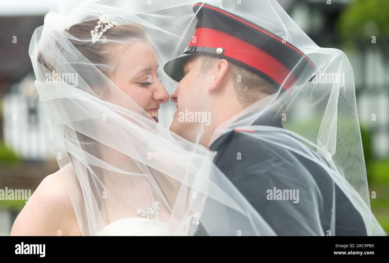 Una giovane sposa bella bacia il sposi marito sotto un velo, dopo aver sposato sua infanzia sweetheart sui loro militari giorno di nozze Foto Stock