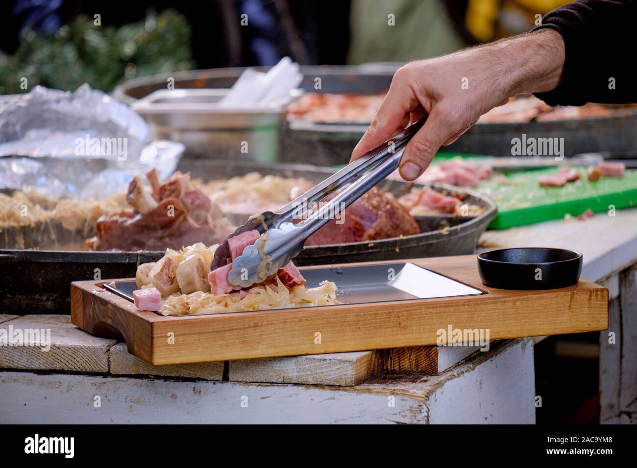 Focus sulla tradizionale croata Buncek- Hock di maiale preparato in una padella grande. Mano di uomo con le tenaglie di maiale di placcatura e il cavolo da cottura preparazione Foto Stock