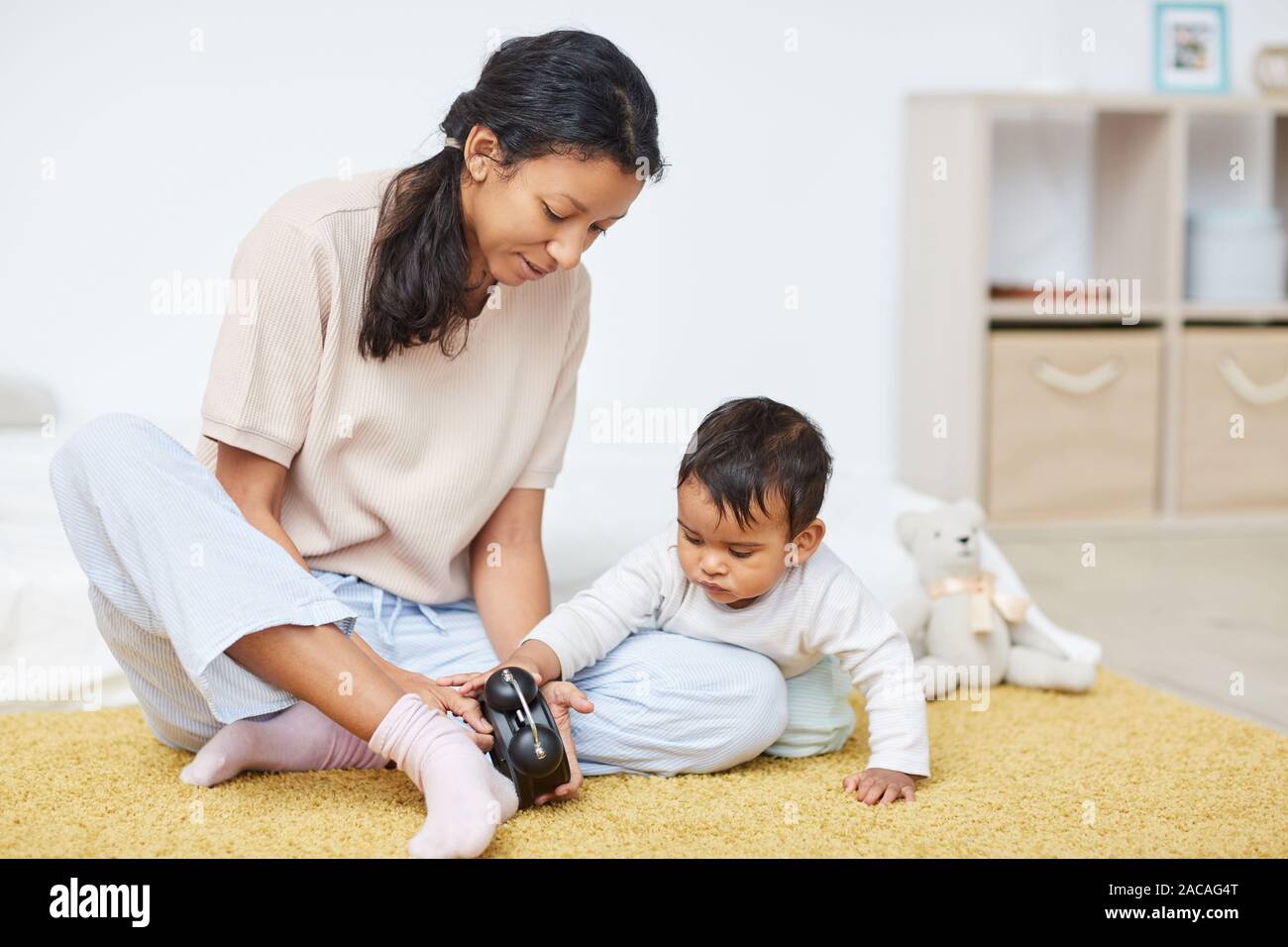 Giovane madre in abbigliamento casual seduto sul pavimento e che mostra il clock per il suo bambino Foto Stock