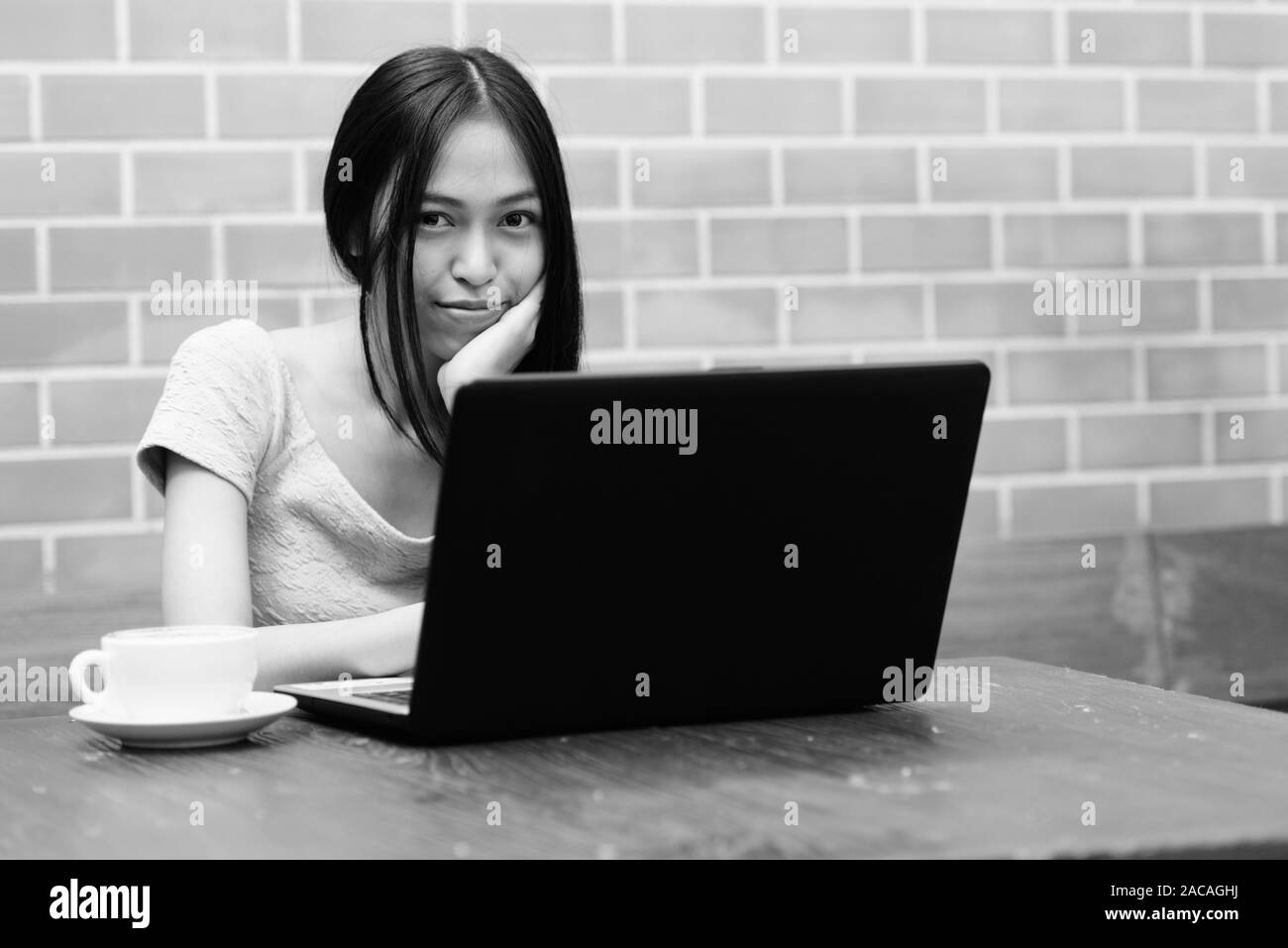 Giovane bella asiatica ragazza adolescente in appoggio il mento sulla mano con laptop e cappuccino sul tavolo di legno contro un muro di mattoni Foto Stock