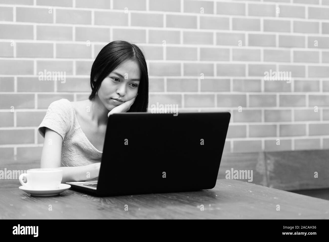 Giovane bella asiatica ragazza adolescente in appoggio il mento sulla mano mentre utilizzando laptop con cappuccino sul tavolo di legno contro un muro di mattoni Foto Stock