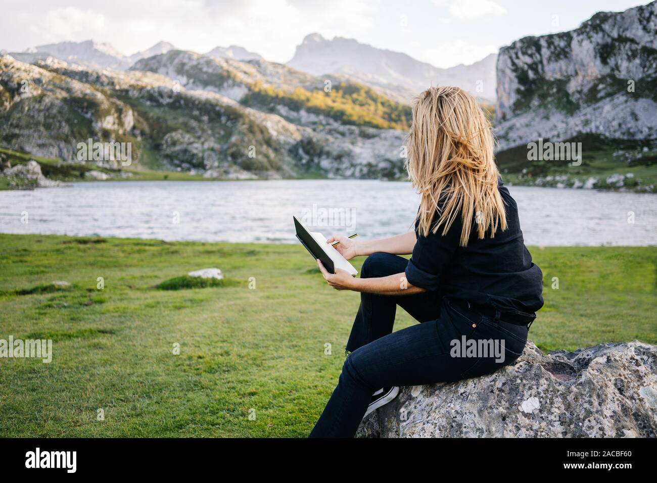 Una bella caucasian donna bionda scrive e disegna su un blocco note in un paesaggio di montagna con il lago Foto Stock