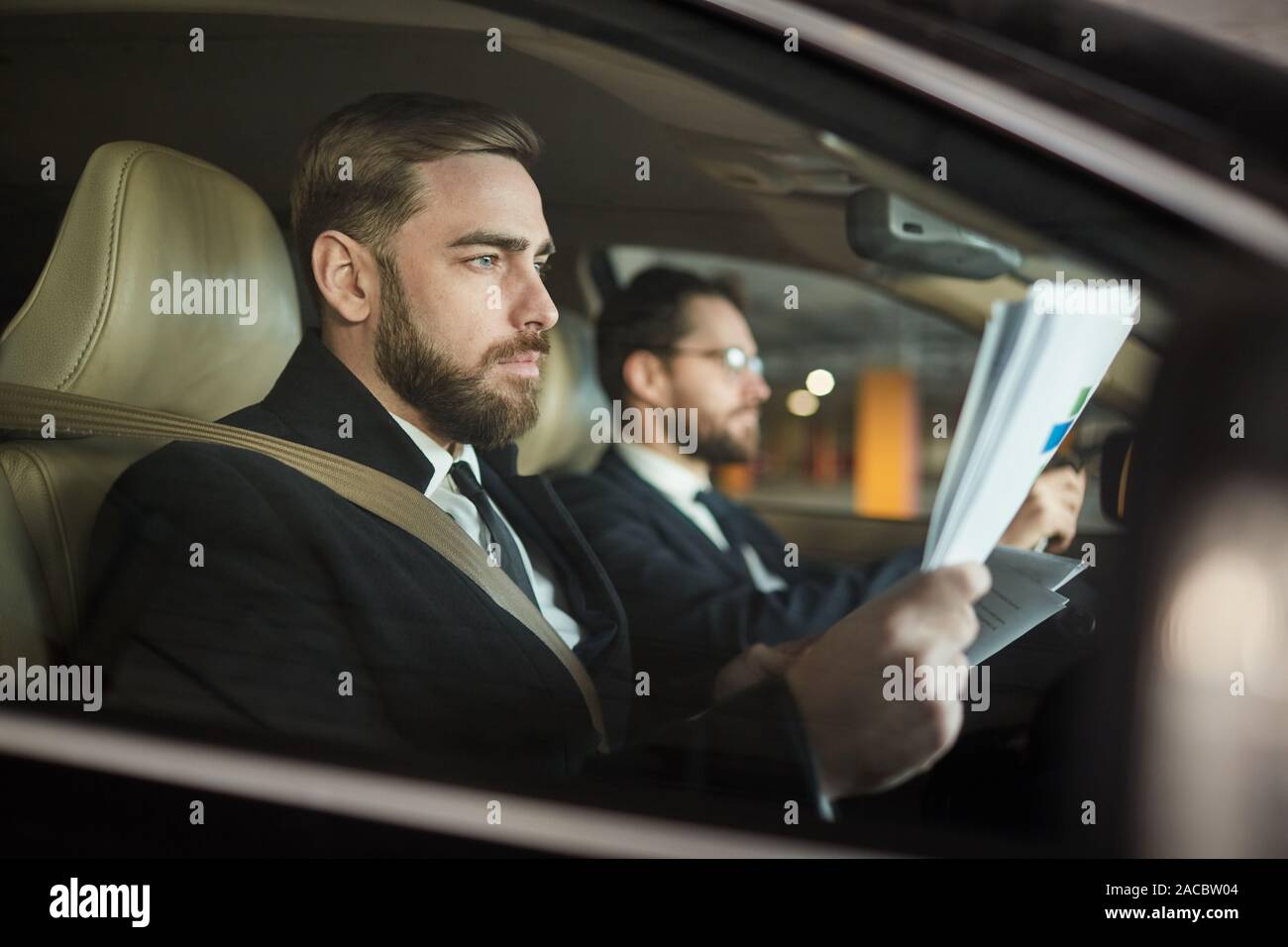 Grave imprenditore barbuto seduto in macchina e la lettura di un contratto con il suo autista alla guida della vettura Foto Stock