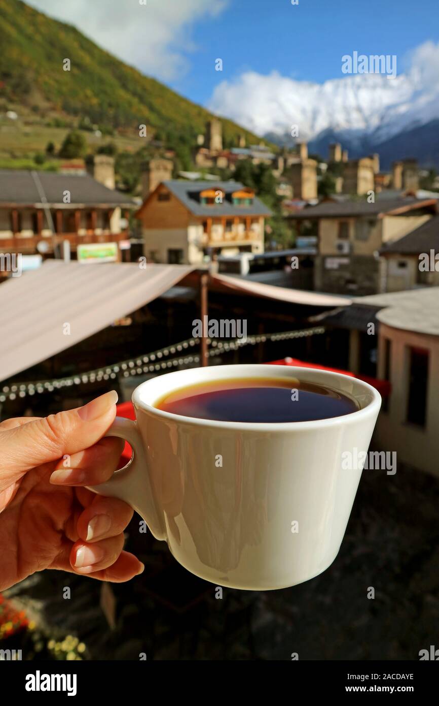 La femmina mano che regge una tazza di caffè caldo contro sfocata montagne del Caucaso e Mestia città in background Foto Stock