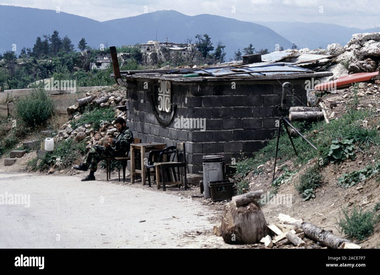 11 agosto 1993 durante l'assedio di Sarajevo: un soldato Bosnian-Serb rilassa fuori del suo bunker sul Monte Trebevic, al di sopra di Sarajevo. Una cinghia di trascorso M2 Browning .50 cartucce calibro appendere al di sotto di un serbo simbolo a croce sulla parte anteriore della capanna. Foto Stock