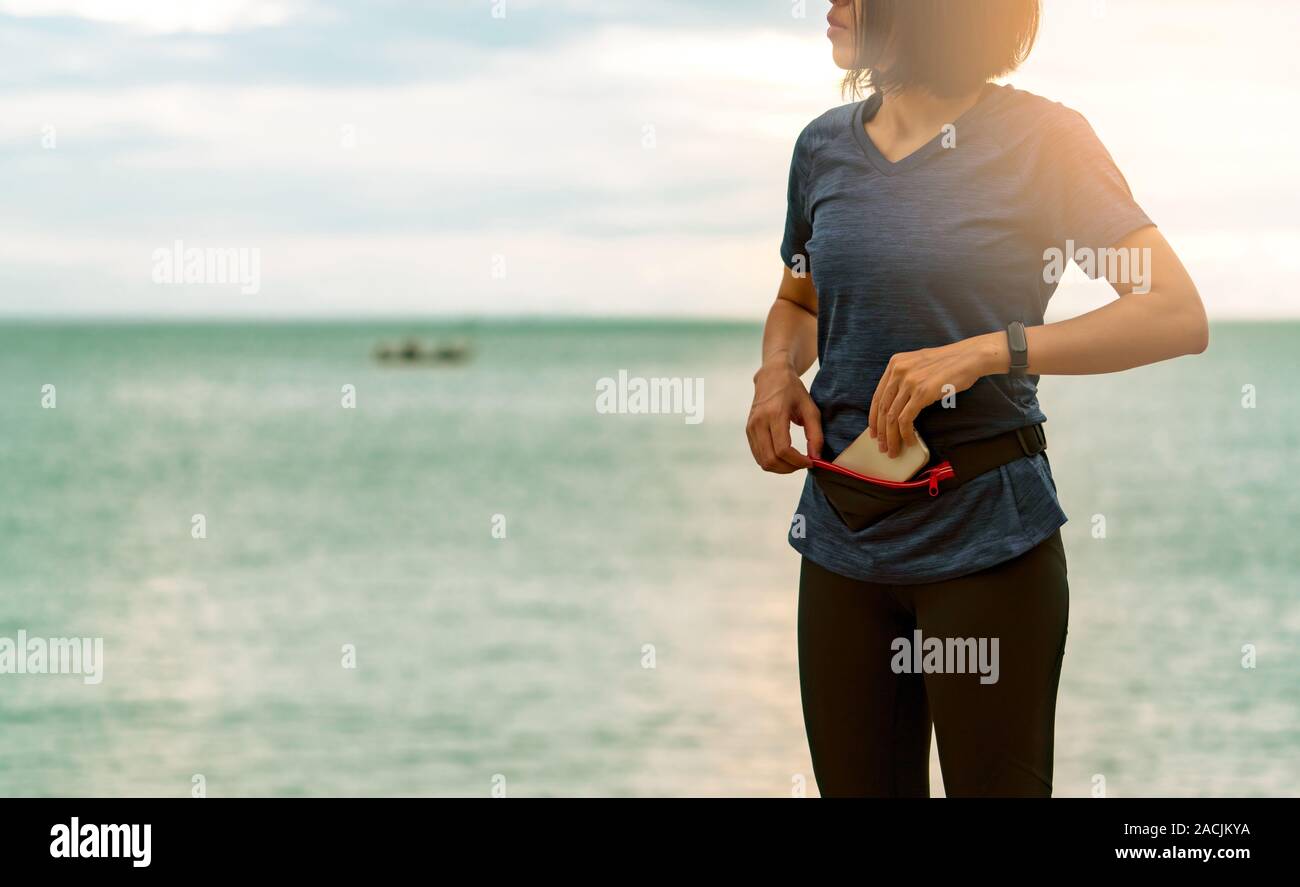 Giovane donna asiatica tenere lo smartphone nel marsupio prima di eseguire cardio esercizio al mattino alla spiaggia del mare. Allenamento all'aperto. Runner e smart usura di banda Foto Stock