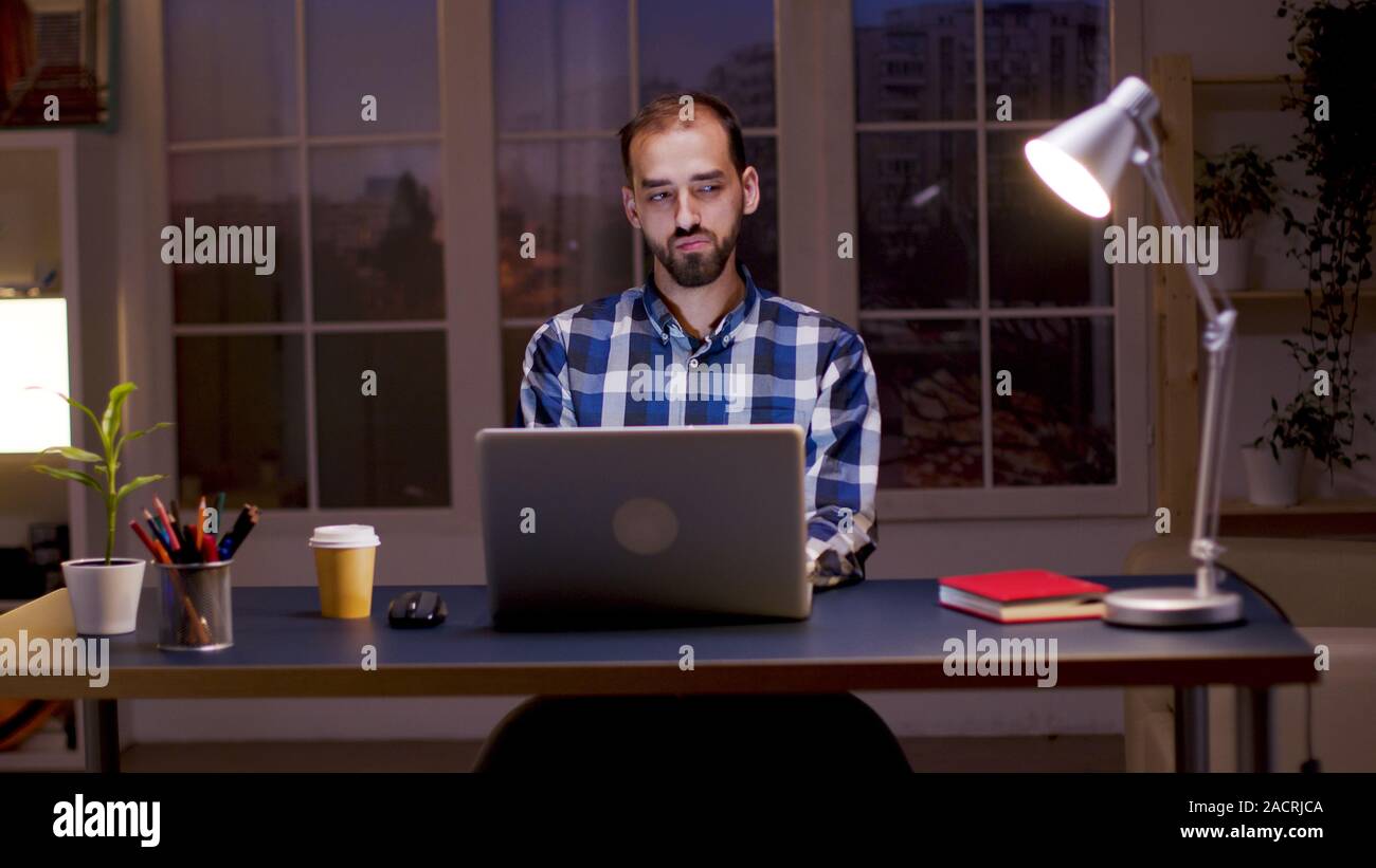 Imprenditore barbuto che indossa una camicia e prendere un sorso di caffè mentre si lavora da casa di notte ore. Foto Stock